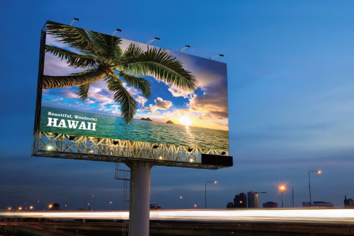 A large raised billboard frontlit at night showing a tropical scene in bright colours
