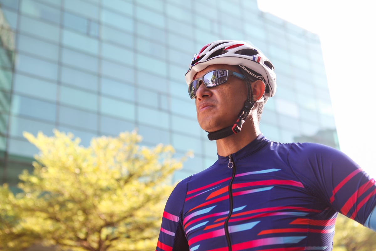 A person wearing a dye sublimation printed cycling jersey with helmet and sunglasses in front of a large glass building
